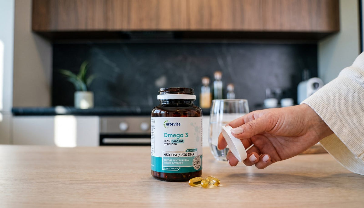 Flacon de Omega 3 Artevita pe blatul bucătăriei, cu capacul alb ținut în mână, un pahar cu apă în spate și două capsule softgel pe lemn.